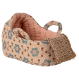A carry cot with a pink floral pattern and teal flowers on a beige background. It features two looped straps and a crocheted front detail, conveying a handcrafted charm.