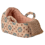 A carry cot with a pink floral pattern and teal flowers on a beige background. It features two looped straps and a crocheted front detail, conveying a handcrafted charm.