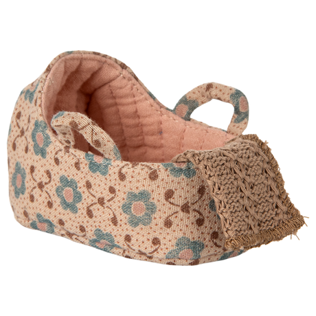 A carry cot with a pink floral pattern and teal flowers on a beige background. It features two looped straps and a crocheted front detail, conveying a handcrafted charm.