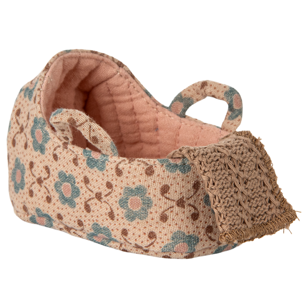 A carry cot with a pink floral pattern and teal flowers on a beige background. It features two looped straps and a crocheted front detail, conveying a handcrafted charm.