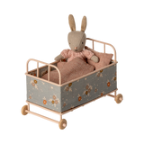 Vintage-style miniature toy crib with floral fabric, soft brown and beige tones. Features small wheels, a houndstooth pillow, and a cozy blanket, evoking a nostalgic charm.