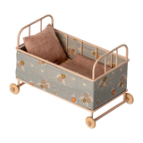Vintage-style miniature toy crib with floral fabric, soft brown and beige tones. Features small wheels, a houndstooth pillow, and a cozy blanket, evoking a nostalgic charm.