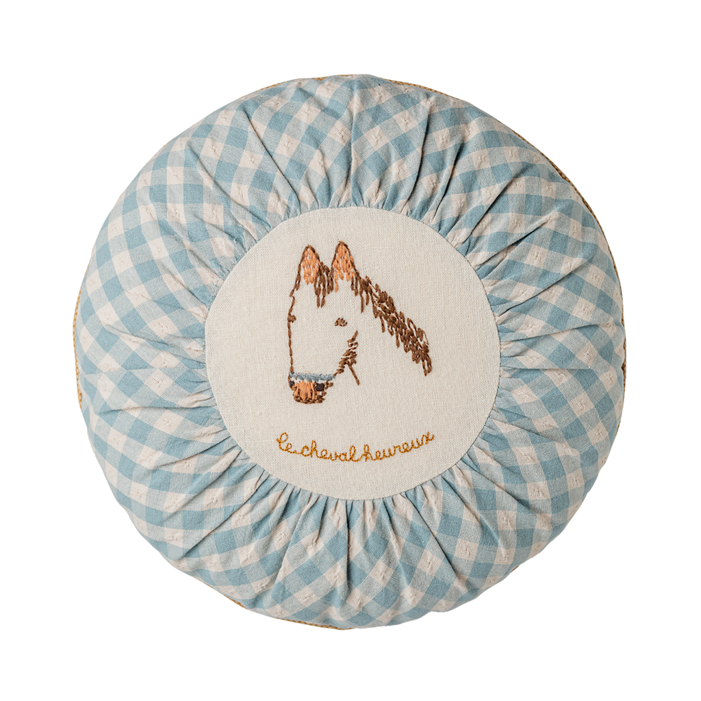 A small, round cushion with a light blue and white gingham pattern around its ruffled edge. The center of the cushion is a light cream circle with an embroidered brown horse head and the words "le cheval heureux" (French for "the happy horse") in a cursive brown script below it.