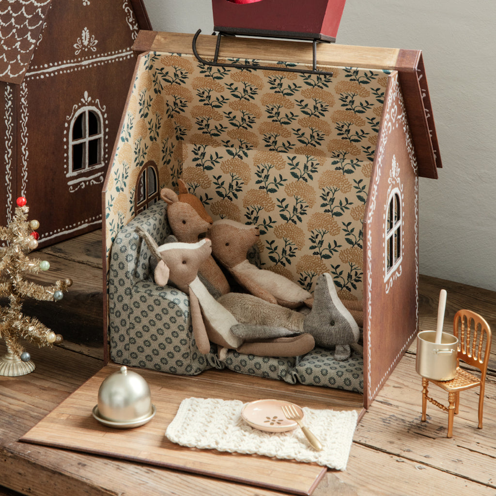 Cozy scene in a dollhouse with toy animals cuddling on a patterned couch. A small gold Christmas tree is nearby, alongside tiny tableware and furniture.