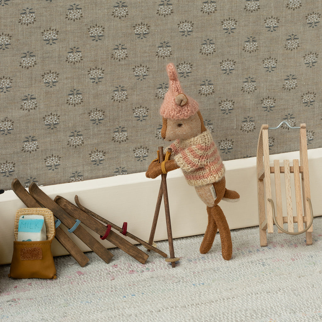 A small, knitted mouse in a sweater and hat leans on ski poles beside wooden skis and a sled. A toy bag with a milk carton is nearby on a patterned carpet.