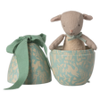 A soft, beige toy rabbit peeks out of a half-open, patterned blue egg-shaped box adorned with white floral designs and a green ribbon, set against a white background.
