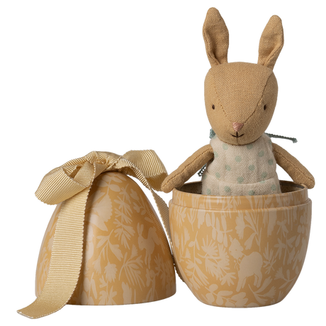 A small, brown plush bunny wearing a polka-dotted outfit sits inside an open, decorative beige egg with floral patterns and a ribbon, against a white background.