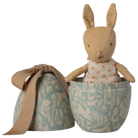 A stuffed bunny in a polka-dot dress peeks out of a decorative blue egg-shaped container with floral patterns and a tied beige ribbon.