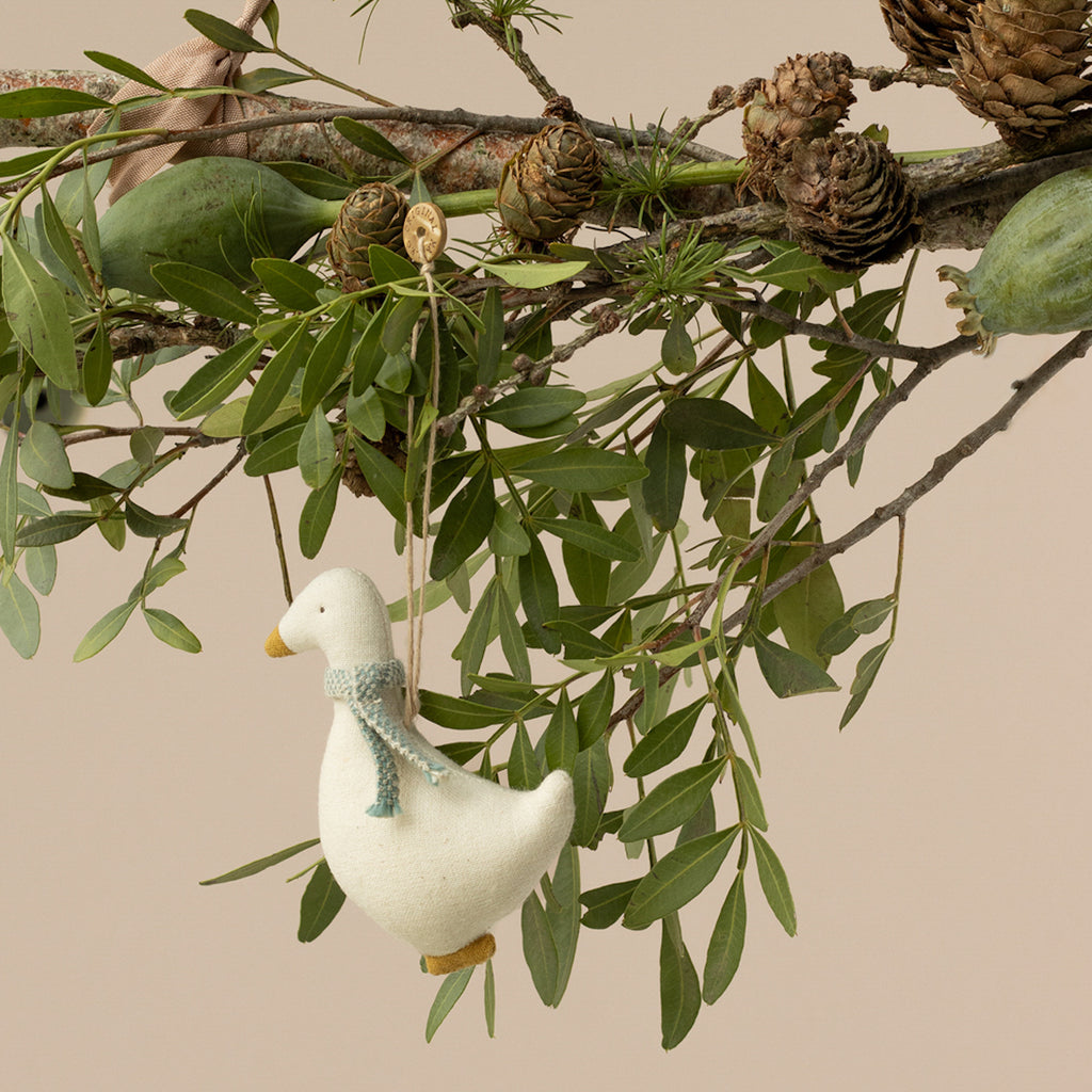 A small, stuffed duck ornament with a blue scarf hangs from a branch adorned with green leaves and pinecones, creating a whimsical, natural scene.