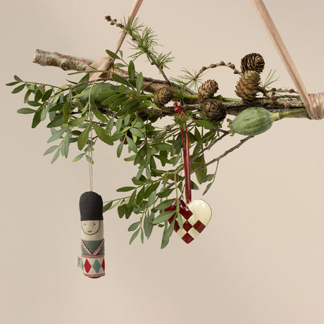 A rustic branch adorned with leafy greenery, pinecones, a toy soldier ornament, and a woven heart dangles on ribbons, evoking a festive, natural ambiance.