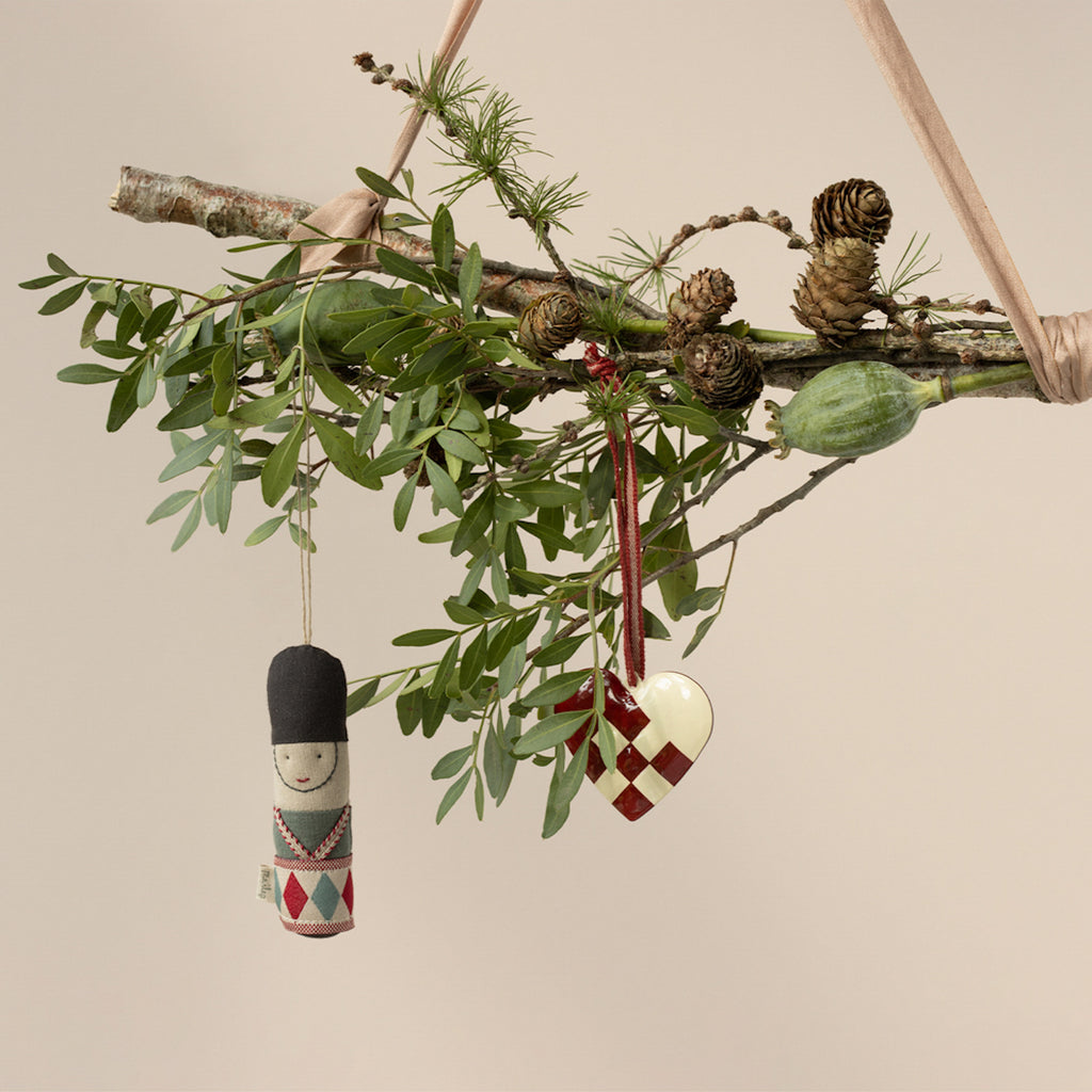 A rustic branch adorned with leafy greenery, pinecones, a toy soldier ornament, and a woven heart dangles on ribbons, evoking a festive, natural ambiance.