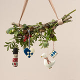 A decorative branch hanging by a ribbon, adorned with greenery, red berries, and small pine cones. Four ornaments hang: a soldier, heart, goose, and drum.