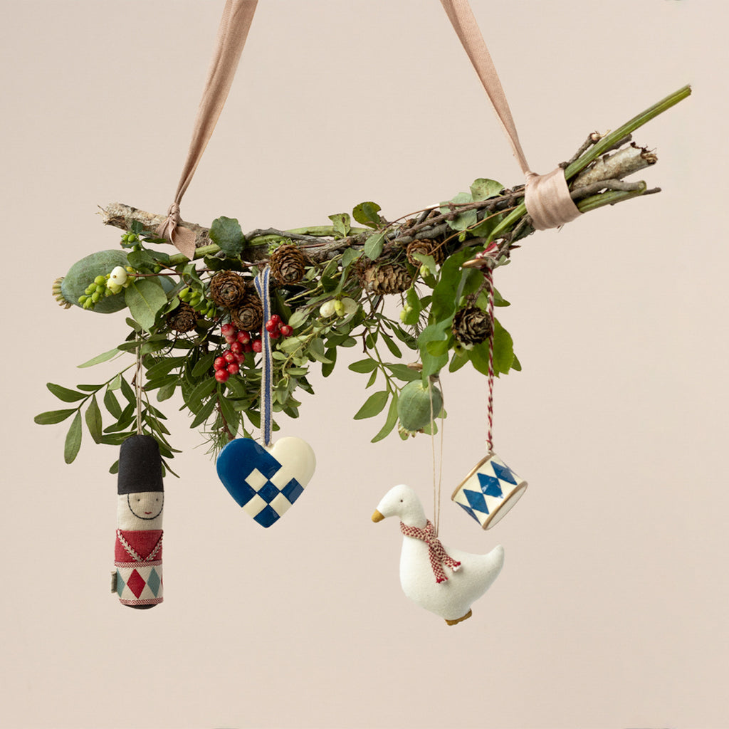 A decorative branch hanging by a ribbon, adorned with greenery, red berries, and small pine cones. Four ornaments hang: a soldier, heart, goose, and drum.