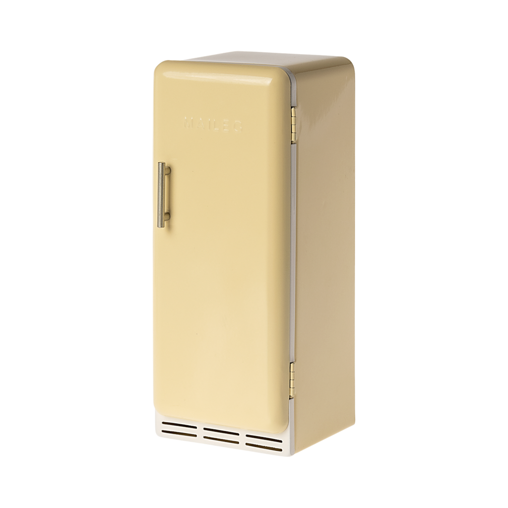 A toy refrigerator in a pale yellow color, is shown with its door slightly ajar. The fridge has a retro design with rounded edges, a silver-colored handle on the right side of the door, and the word "MAILEG" embossed vertically on the door. Inside, three shelves with ribbed textures are visible.