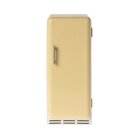 A toy refrigerator in a pale yellow color, is shown with its door slightly ajar. The fridge has a retro design with rounded edges, a silver-colored handle on the right side of the door, and the word "MAILEG" embossed vertically on the door. Inside, three shelves with ribbed textures are visible.