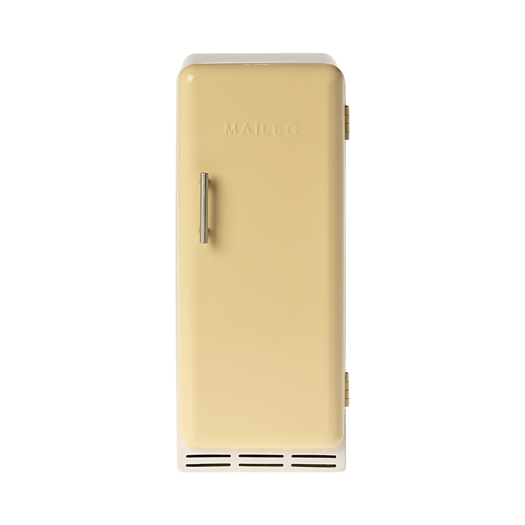 A toy refrigerator in a pale yellow color, is shown with its door slightly ajar. The fridge has a retro design with rounded edges, a silver-colored handle on the right side of the door, and the word "MAILEG" embossed vertically on the door. Inside, three shelves with ribbed textures are visible.