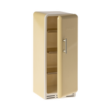 A toy refrigerator in a pale yellow color, is shown with its door slightly ajar. The fridge has a retro design with rounded edges, a silver-colored handle on the right side of the door, and the word "MAILEG" embossed vertically on the door. Inside, three shelves with ribbed textures are visible.