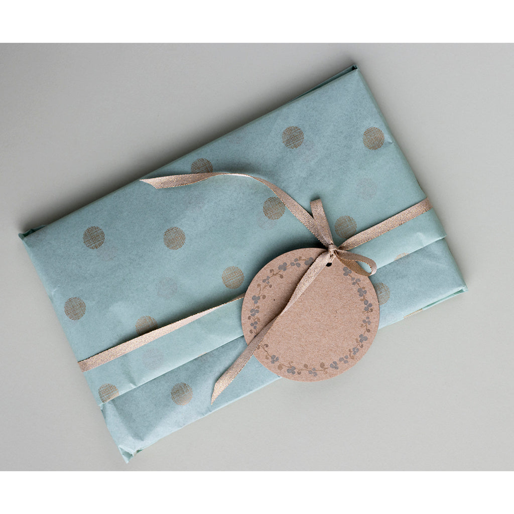 A gift wrapped in light blue polka dot paper with a beige ribbon and a round brown tag attached, lying on a grey surface. The tone is elegant and festive.