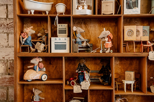 Wooden display shelving filled with Maileg mouse figures and miniature furniture, including a bath, cooker, bicycles and alarm clocks