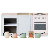 Miniature toy kitchen set with a pink countertop, mini sink, stovetop, and oven. In front are colorful cups, a can of tomatoes, and a small saucepan. Charming and playful.