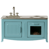 A miniature blue kitchen set with a sink, faucet, and stovetop. An oven with a glass door shows a pie inside. The tone is playful and nostalgic.