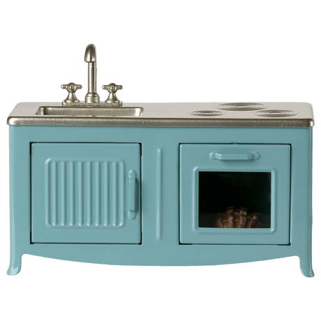 A miniature blue kitchen set with a sink, faucet, and stovetop. An oven with a glass door shows a pie inside. The tone is playful and nostalgic.