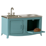 A miniature blue kitchen with a silver sink and stove. The open oven door reveals a small loaf of bread. The scene conveys a playful, retro vibe.