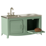 A mint-green toy kitchen set with a small sink, burner marks, and an open oven door revealing a miniature pie. The scene evokes playful nostalgia.
