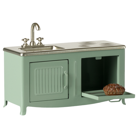 A mint-green toy kitchen set with a small sink, burner marks, and an open oven door revealing a miniature pie. The scene evokes playful nostalgia.
