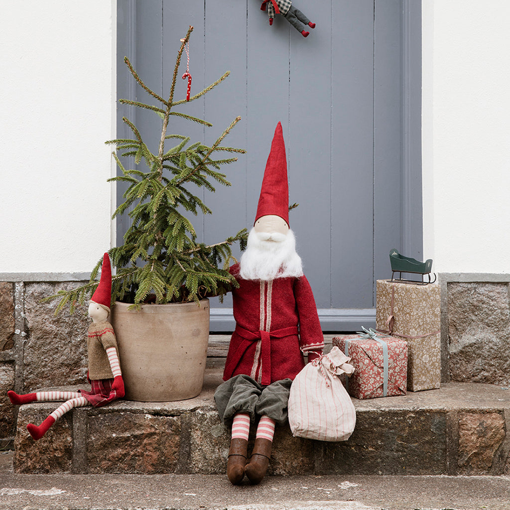 Festive scene with two red-hatted gnomes, a small pine tree, and wrapped gifts on a stone doorstep. Cozy, holiday atmosphere.