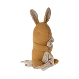 A soft, brown stuffed bunny with patterned fabric ears sits holding a white, polka-dotted blanket. The cozy scene conveys warmth and comfort.