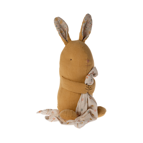 A soft, brown stuffed bunny with patterned fabric ears sits holding a white, polka-dotted blanket. The cozy scene conveys warmth and comfort.