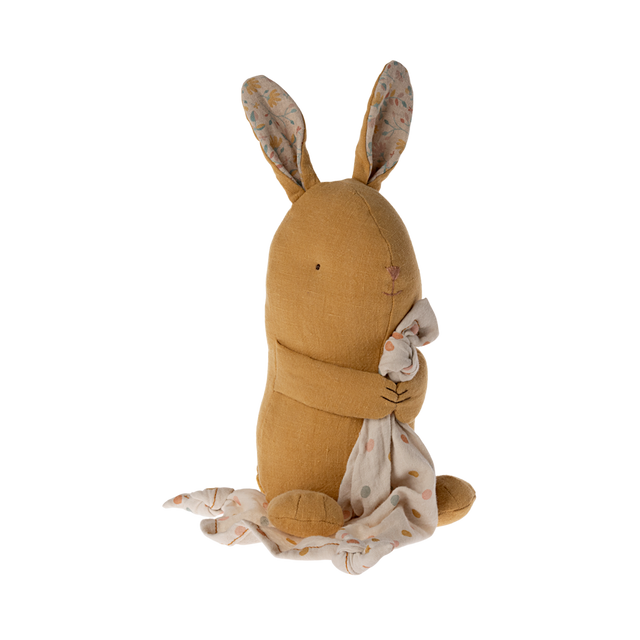 A soft, brown stuffed bunny with patterned fabric ears sits holding a white, polka-dotted blanket. The cozy scene conveys warmth and comfort.