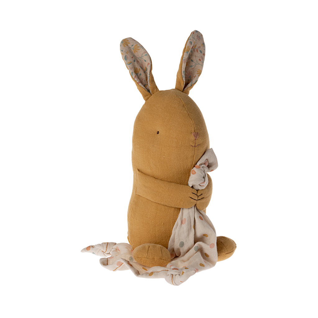 A soft, brown stuffed bunny with patterned fabric ears sits holding a white, polka-dotted blanket. The cozy scene conveys warmth and comfort.