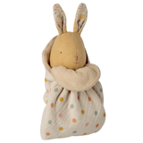 A soft toy rabbit with embroidered features and patterned ears is wrapped in a cream polka dot fabric. The tone is cozy and whimsical.