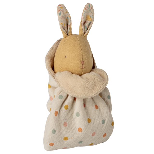 A soft toy rabbit with embroidered features and patterned ears is wrapped in a cream polka dot fabric. The tone is cozy and whimsical.