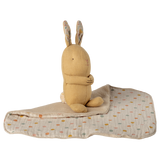 A soft, beige stuffed rabbit with folded arms and patterned ears sits on a quilt with pastel polka dots. The background is white, creating a cozy, serene feel.