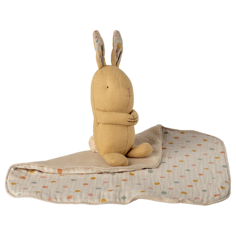 A soft, beige stuffed rabbit with folded arms and patterned ears sits on a quilt with pastel polka dots. The background is white, creating a cozy, serene feel.
