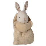 A plush toy rabbit with floral-patterned ears and an embroidered face is snugly swaddled in a soft, polka-dotted beige blanket, set against a white background.