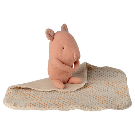 A soft pink stuffed animal resembling a small rodent sits on a beige blanket with brown polka dots, set against a white background. The tone is cozy.