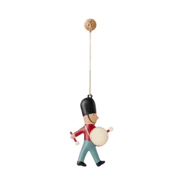 A hanging ornament of a toy soldier wearing a tall black hat, red jacket, and blue pants, playing a drum. The toy is suspended by a string, conveying a playful and cheerful tone.
