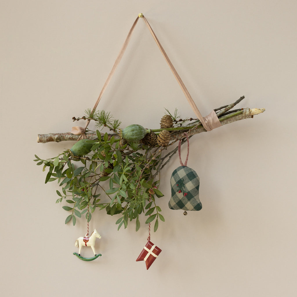 Festive wall decoration with branches, green leaves, pinecones, and ornaments: a rocking horse, plaid bell, and red gift. Earthy, rustic theme.