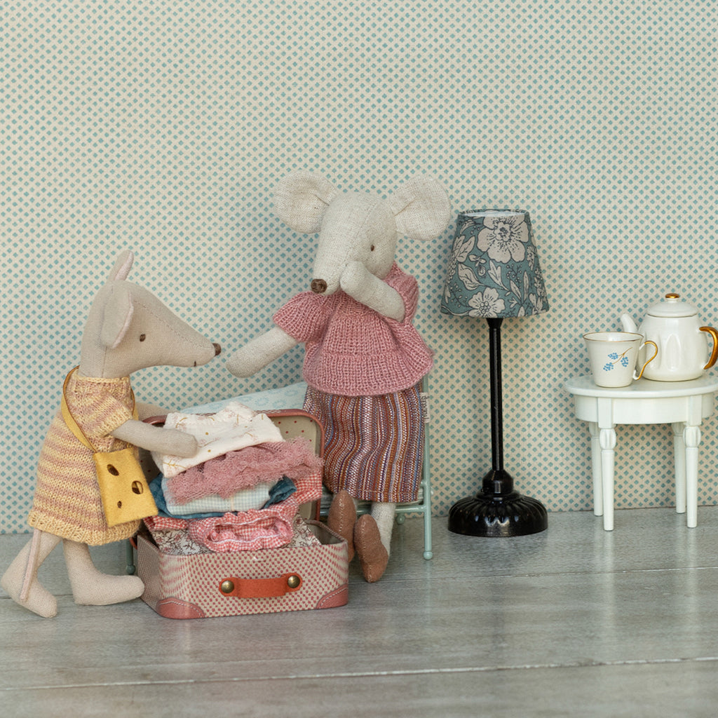 Two fabric mice, one in yellow, one in pink, pack a small suitcase. A lamp and teapot sit nearby. The scene is cozy and playful.