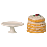 White cake stand next to a stack of pancakes topped with cream and berries, set against a white background. The scene feels whimsical and playful.