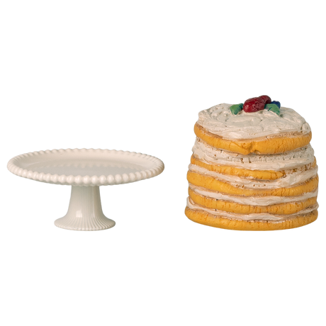 White cake stand next to a stack of pancakes topped with cream and berries, set against a white background. The scene feels whimsical and playful.