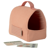 A soft, peach-colored pet carrier with a red grid pattern and handle. Nearby are a Pet Journal and dog stickers against a white background, conveying a cozy, playful vibe.