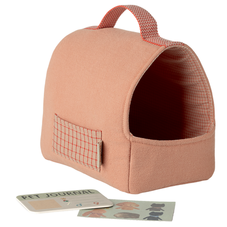 A soft, peach-colored pet carrier with a red grid pattern and handle. Nearby are a Pet Journal and dog stickers against a white background, conveying a cozy, playful vibe.