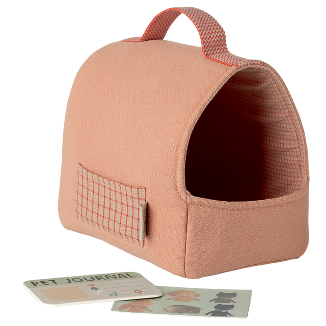 A soft, peach-colored pet carrier with a red grid pattern and handle. Nearby are a Pet Journal and dog stickers against a white background, conveying a cozy, playful vibe.