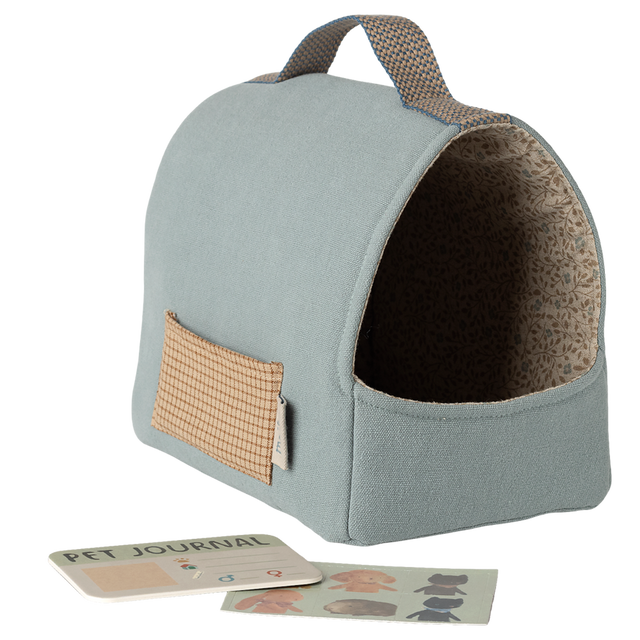 Light blue fabric pet carrier with a brown patterned interior and a small external pocket. A pet journal and stickers lie in front, all set against a white background.
