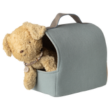 A fluffy brown teddy bear peeks out of a light blue, fabric carrier with a handle. The setting is whimsical and cute against a white background.